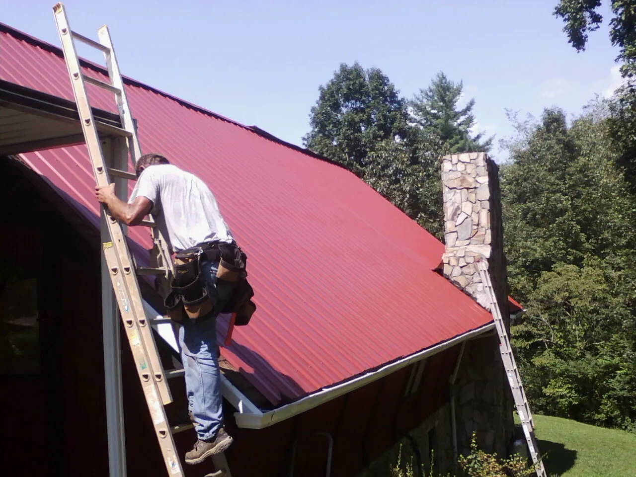 Roofer on a ladder beside a red metal roof and stone chimney — Soffit & Fascia Repair by Roofing Brothers in Whiting